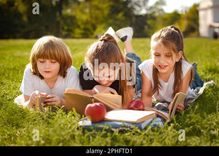 I bambini si posano in fila su erba verde e leggono libri nel parco dopo le lezioni scolastiche Foto Stock