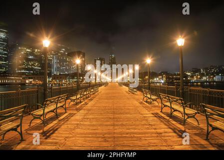 Deserto legno Pier sette fiancheggiato da luci di strada e panchine a San Francisco di notte. Lo skyline del centro cittadino parzialmente coperto di nebbia e' sullo sfondo. Foto Stock