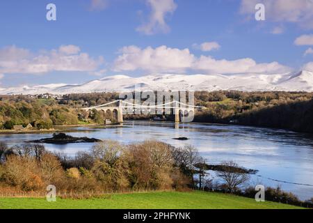 Vista panoramica sul ponte sospeso di Menai che attraversa lo stretto di Menai con neve sulle montagne in inverno. Menai Bridge, Isola di Anglesey, Galles, Regno Unito Foto Stock