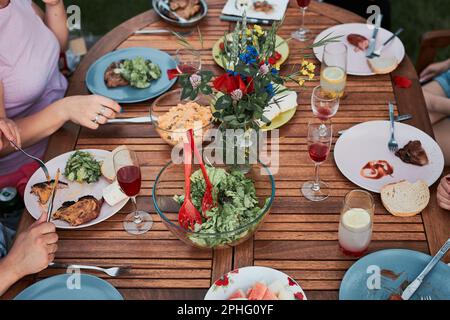 Famiglia che ha un pasto dalla griglia durante la cena estiva picnic all'aperto in un giardino di casa. Primo piano di persone sedute a un tavolo con cibo e piatti Foto Stock