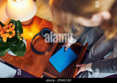 Giovane donna che tira l'agenda dal cassetto del comodino. Scrittura giornale. Creazione di note personali Foto Stock