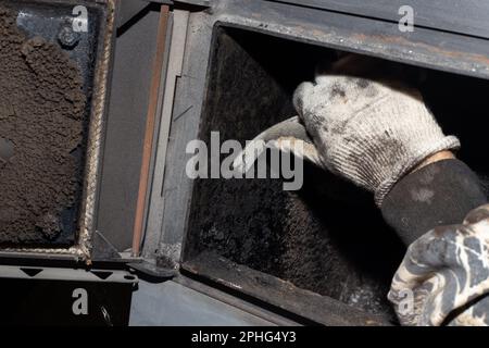 Un uomo pulisce una caldaia a combustibile solido da fuliggine e fuliggine. Manutenzione dell'impianto di riscaldamento dopo la stagione di riscaldamento. Foto Stock