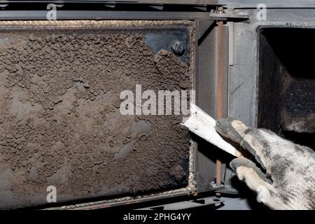 Un uomo pulisce una caldaia a combustibile solido da fuliggine e fuliggine. Manutenzione dell'impianto di riscaldamento dopo la stagione di riscaldamento. Foto Stock