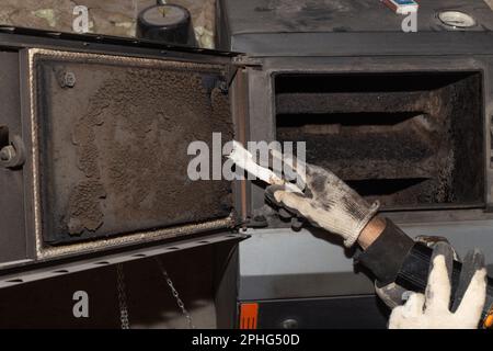 Un uomo pulisce una caldaia a combustibile solido da fuliggine e fuliggine. Manutenzione dell'impianto di riscaldamento dopo la stagione di riscaldamento. Foto Stock