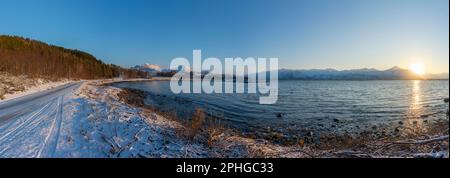 Abendrot mit einem hellen Sonnenstrahl über den verschneiten von Senja, Norwegen. Sandstrand im Winter am Ufer des Fjords bei Skaland, Troms Foto Stock