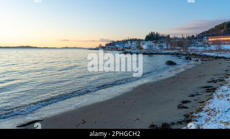 Abendrot mit einem hellen Sonnenstrahl über den verschneiten von Senja, Norwegen. Sandstrand im Winter am Ufer des Fjords bei Skaland, Troms Foto Stock