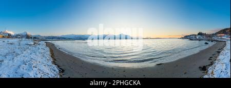 Abendrot mit einem hellen Sonnenstrahl über den verschneiten von Senja, Norwegen. Sandstrand im Winter am Ufer des Fjords bei Skaland, Troms Foto Stock