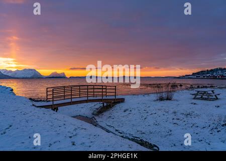 Abendrot mit Brücke und einem hellen Sonnenstrahl über verschneiten von Senja, Norwegen. Die roten Wolken spiegeln sich im Fjord bei Skaland Foto Stock