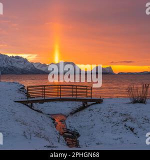 Abendrot mit Brücke und einem hellen Sonnenstrahl über verschneiten von Senja, Norwegen. Die roten Wolken spiegeln sich im Fjord bei Skaland Foto Stock