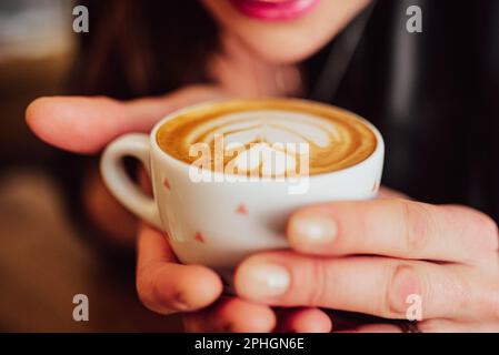 le mani che tengono una tazza bianca con caffè e latte art in un caffè, una donna e labbra dipinte possono essere viste sullo sfondo Foto Stock