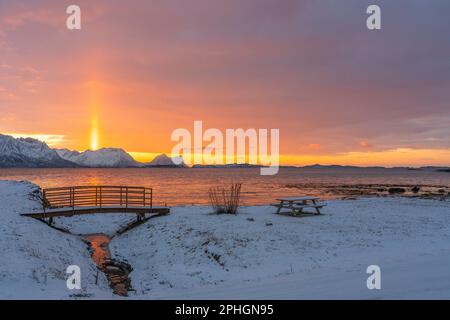 Abendrot mit Brücke und einem hellen Sonnenstrahl über verschneiten von Senja, Norwegen. Die roten Wolken spiegeln sich im Fjord bei Skaland Foto Stock