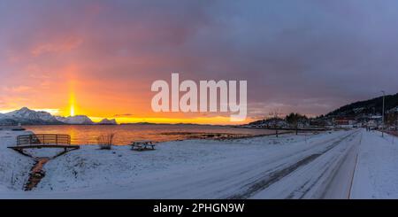 Abendrot mit Brücke und einem hellen Sonnenstrahl über verschneiten von Senja, Norwegen. Die roten Wolken spiegeln sich im Fjord bei Skaland Foto Stock