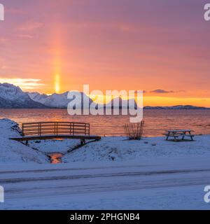 Abendrot mit Brücke und einem hellen Sonnenstrahl über verschneiten von Senja, Norwegen. Die roten Wolken spiegeln sich im Fjord bei Skaland Foto Stock