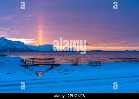 Abendrot mit Brücke und einem hellen Sonnenstrahl über verschneiten von Senja, Norwegen. Die roten Wolken spiegeln sich im Fjord bei Skaland Foto Stock