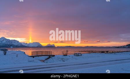 Abendrot mit Brücke und einem hellen Sonnenstrahl über verschneiten von Senja, Norwegen. Die roten Wolken spiegeln sich im Fjord bei Skaland Foto Stock