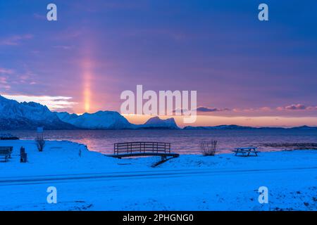 Abendrot mit Brücke und einem hellen Sonnenstrahl über verschneiten von Senja, Norwegen. Die roten Wolken spiegeln sich im Fjord bei Skaland Foto Stock