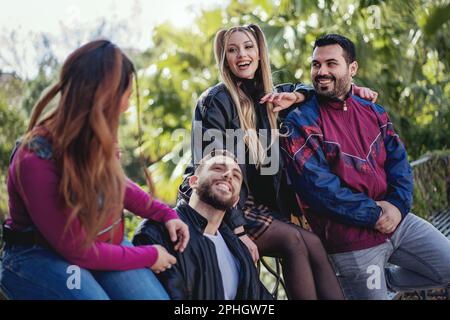 Gruppo di quattro giovani adulti felici, due uomini e due donne, chiacchierando e sorridendo su una panchina del parco in una giornata di sole, indossando abiti vintage 90s, circondati Foto Stock