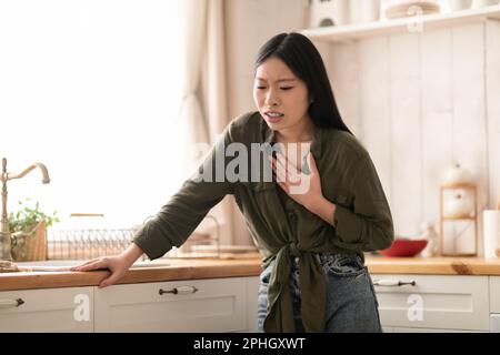 Donna cinese malata che soffre di bruciore di stomaco in cucina, toccando il petto Foto Stock