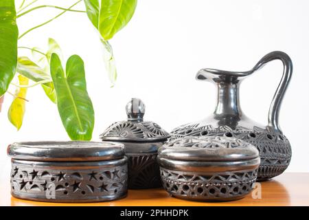 Artigianato tipico messicano. Set di vasi di argilla nera fatti a mano a Oaxaca, Messico. Fondo bianco, base in legno e pianta sullo sfondo. Foto Stock