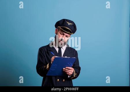 Pilota di aeroplano che tiene appunti, scrivendo, aviatore concentrato con documenti. Capitano di velivolo in uniforme professionale modulo di riempimento in vista frontale dell'aeroporto, studio medio girato su sfondo blu Foto Stock