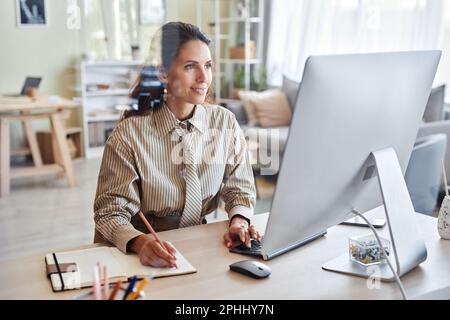 Ritratto di donna sorridente che lavora con il computer alla scrivania dietro una parete di vetro in un ambiente accogliente Foto Stock