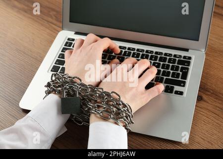 Donna con le mani incatenate che scrivono sul laptop al tavolo di legno, primo piano. Dipendenza da Internet Foto Stock