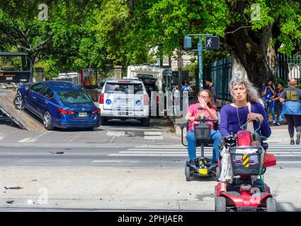 NEW ORLEANS, LA, USA - 26 MARZO 2023: Paesaggio urbano con due donne con una disabilità sui loro scooter motorizzati in un crosswalk e un'automobile che è trainata Foto Stock