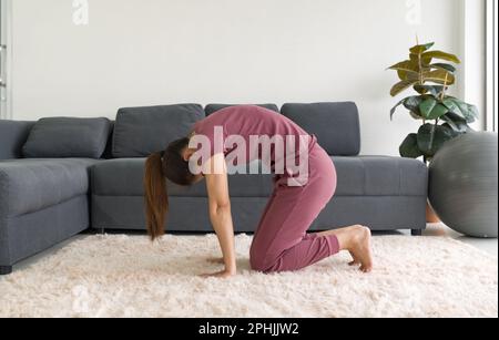 Casalinga asiatica che fa l'esercitazione di posa del gatto durante l'esercitazione domestica. Donna che pratica yoga in Marjaryasana posa sul tappeto nel soggiorno. Foto Stock