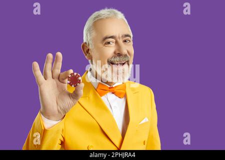 Felice uomo anziano in tuta gialla che si diverte al casinò, sorridendo e mostrando chip di poker Foto Stock