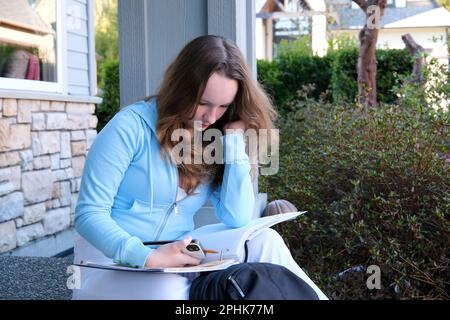 ragazza adolescente fa lavoro sul portico di casa scrive con la matita in notebook grande in cartella accanto allo zaino primavera lavoro sedersi sulla soglia sul portico donna scrive disegna risolve i problemi Foto Stock