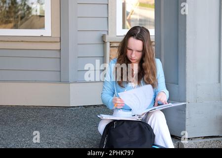 ragazza adolescente fa lavoro sul portico di casa scrive con la matita in notebook grande in cartella accanto allo zaino primavera lavoro sedersi sulla soglia sul portico donna scrive disegna risolve i problemi Foto Stock