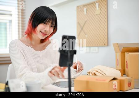 Sorridente e allegro giovane asiatico donna negozio online proprietario che vende i suoi prodotti attraverso live streaming sulla piattaforma di shopping, lavorando da casa. Foto Stock