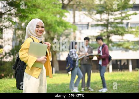 Giovane studentessa asiatica musulmana allettante e sorridente in hijab che porta il suo laptop e zaino e si trova nel parco del campus. Foto Stock