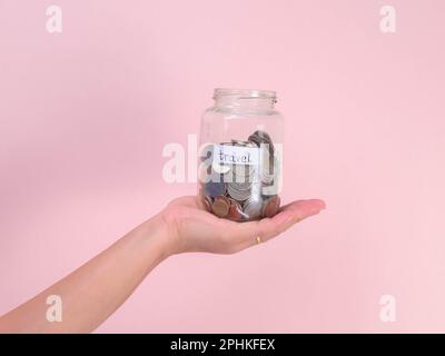 Primo piano della mano della giovane donna che tiene il vaso di vetro con soldi (moneta) all'interno su sfondo rosa. Mano che tiene un vaso di soldi, viaggi, risparmi, istruzione, fare Foto Stock