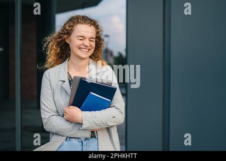 Ritratto di una studentessa che si trova fuori dal campus universitario e che indossa una giacca, un libro e un computer portatile con pausa caffè dopo una lezione. Godendo Foto Stock