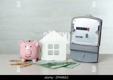 Contatore elettrico, banco di porcellini, modello di casa e banconote in euro su tavolo di legno bianco Foto Stock