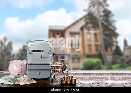 Contatore elettrico, banca di piggy e piccolo carrello della spesa con soldi sul tavolo di legno contro la vista sfocata della bella casa Foto Stock