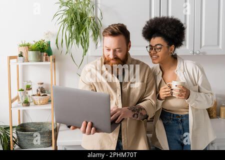 Tatuato uomo utilizzando laptop vicino afroamericano ragazza con tazza in cucina, immagine stock Foto Stock