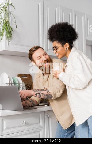 Tatuato uomo utilizzando il laptop e guardando ragazza afro-americana con caffè in cucina, immagine stock Foto Stock