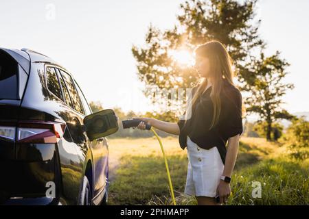 Giovane donna che collega il caricabatterie in un'auto elettrica nera, concetto di energia rinnovabile Foto Stock