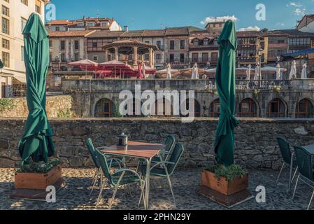 Terrazza con i suoi tavoli, sedie e ombrelloni sulla parete di pietra del fiume con la vista delle case e del tempio sullo sfondo, Potes, Foto Stock