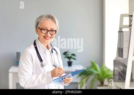 Felice medico senior con appunti in piedi in ufficio medico e sorridente Foto Stock