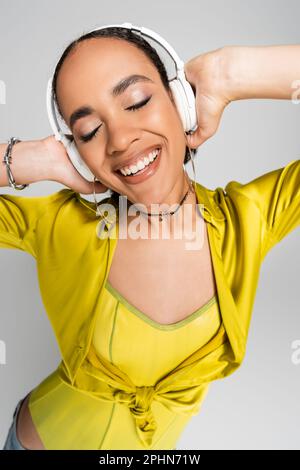 felice afroamericana donna ascoltare musica in cuffie wireless isolato su grigio, immagine stock Foto Stock