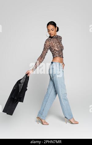 intera lunghezza di modello afro-americano ben vestito in crop top con stampa animale tenendo giacca nera e camminare su grigio, immagine stock Foto Stock