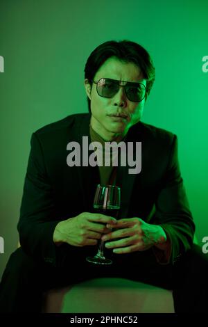 Accigliante uomo asiatico in occhiali da sole con bicchiere di vino rosso Foto Stock