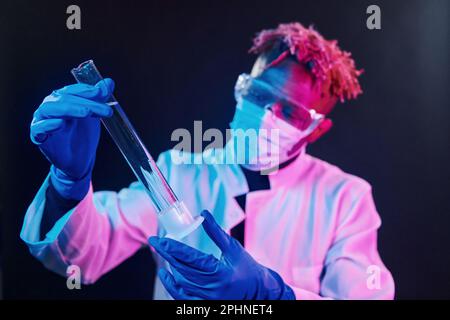 Scienziato intelligente in provetta di tenuta protettiva uniforme. Concezione del coronavirus. Illuminazione futuristica al neon. Giovane afro-americano in studio Foto Stock