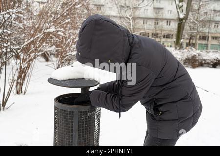 Homeless Man rummages in un cestino può, raccogliendo bottiglie vuote, Beggar alla ricerca di cibo a Cold Winter City, problema di disoccupazione Foto Stock