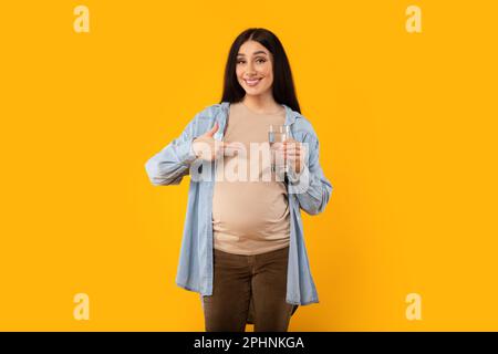 Felice donna incinta che tiene un bicchiere d'acqua e che indica, sorridendo alla macchina fotografica su sfondo giallo studio Foto Stock