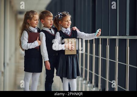 Bambini in uniforme con telefono e selfie in corridoio. Concezione dell'istruzione Foto Stock