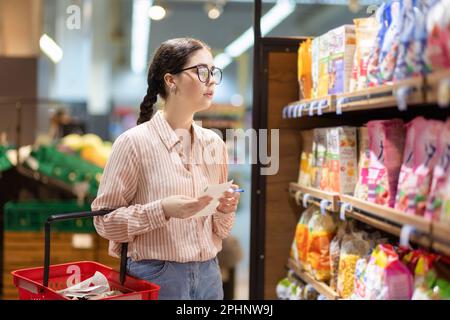 Vista laterale di giovane donna caucasica graziosa che indossa gli occhiali contiene la lista dei prodotti e scrivere in esso. Concetto di shopping in supermercato e consumismo. Foto Stock